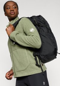Rucksack mit schwarzem Stoffäußeren, mehreren Riemen und Taschen. Kombiniert mit einer grünen Fleecejacke, die ein Logo-Patch zeigt.