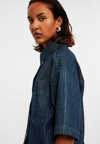 Denim overhemd in donkerblauwe visgraatprint, korte mouwen en een kraag. Met een borstzak en subtiele stikdetails.