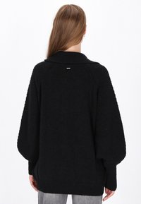 Svart oversized tröja med en texturerad baksida, ribbade ärmslut och en hög krage. Har en discret silverlogotyp på baksidan.