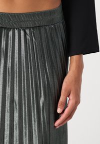 Gonna plissettata grigio scuro realizzata in tessuto lucido con vita elasticizzata. Primo piano di una mano appoggiata accanto alla gonna, che mostra la trama e le pieghe.