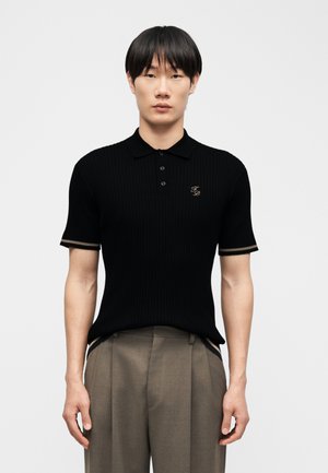 Νεαρός άνδρας με ίσια μαύρα μαλλιά, που φοράει μαύρο κοντομάνικο πόλο με κεντημένα αρχικά και άνετο μπεζ παντελόνι.