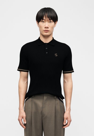 Jeune homme aux cheveux noirs et raides portant un polo noir à manches courtes avec des initiales brodées et un pantalon beige ample.
