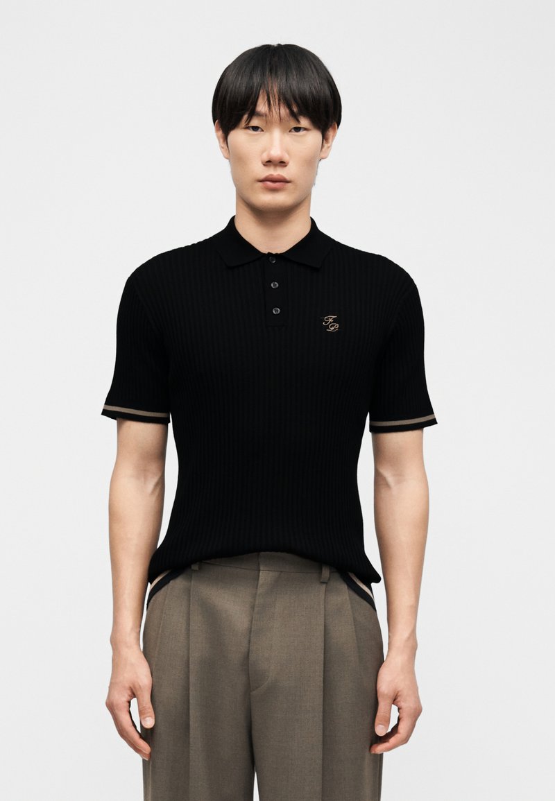 Noor mees sirgete mustade juustega, kandes musta lühikeste varrukatega polot, millel on tikitud initsiaalid, ning laiu beeže pükse.