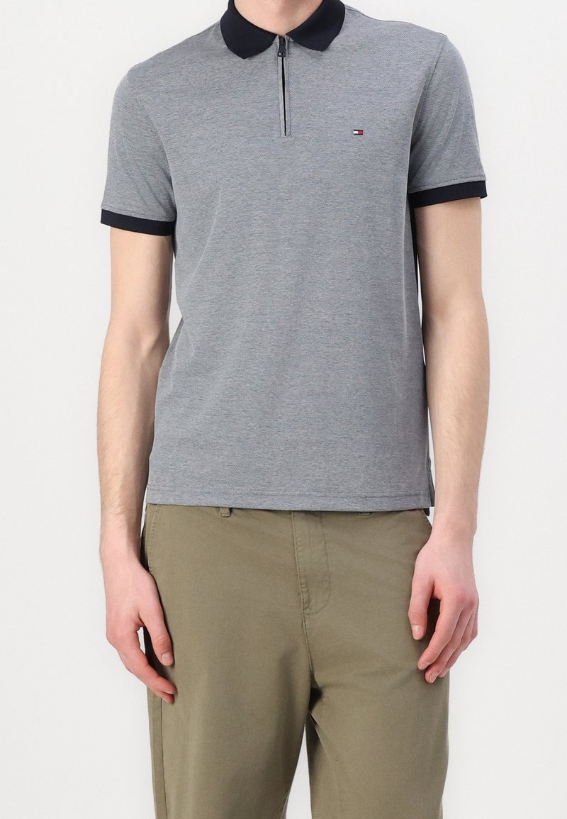 Polo shirt gris à manches courtes avec un col bleu marine foncé et un devant zippé, arborant un petit logo sur la poitrine gauche, fabriqué à partir d'un tissu texturé.