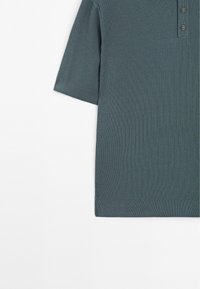 Koszulka polo z krótkim rękawem w ciemnym odcieniu zieleni morska, o prążkowanej fakturze, z trzema guzikami i subtelnymi detalami szwów.