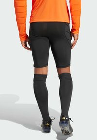 Schwarze sportliche Shorts mit enger Passform und Seitentaschen. Getragen mit kniehohen schwarzen Socken und Stollen mit goldenen Akzenten. Orangenes Langarmshirt.