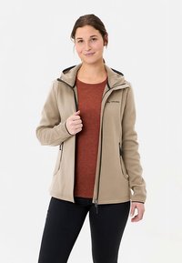 Beige Jacke zum Reißverschluss mit Kapuze, kontrastierenden schwarzen Akzenten, Seitentaschen und weicher Textur, getragen über einem rostfarbenen Oberteil.