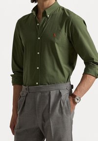 Donkergroen overhemd met knopen en een kraag, met een klein rood embleem. Gecombineerd met grijze geplooide broek en een bruine horloge.