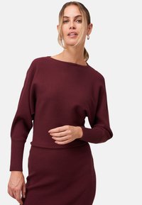 Pull en rib d'un couleur bordeaux avec un décolleté bateau et des manches longues, associé à une jupe en rib assortie. Le tissu a une texture douce.