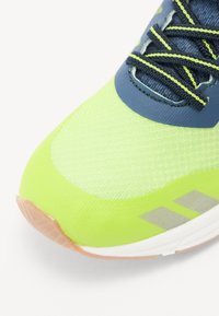Neon zielona cholewka sportowego buta z teksturowaną siatką, niebieskimi akcentami i odbijającymi szarymi paskami. Gumowa podeszwa w beżowym odcieniu. Sznurówki w czarnym kolorze z neonowymi krawędziami.