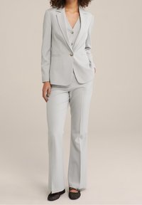 Ljust grå skräddarsydd kostym med spetsigt krage, tvåknappsstängning och utsvängda byxor. Slätt tyg med en elegant design. Svarta platta skor.