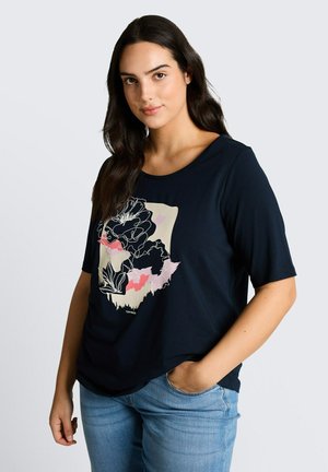 Kobieta z długimi ciemnymi włosami nosi granatową koszulkę z kwiatowym nadrukiem i niebieskie jeansy, pozując z jedną ręką w kieszeni na jasnym tle.