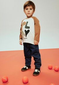 Long-sleeve white shirt with brown sleeves featuring "PLAY" graphic, paired with blue jeans and black sneakers with white accents, standing on orange background.
