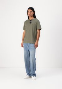 T-shirt in cotone verde oliva abbinato a jeans loose-fit azzurri chiari. Modello con occhiali da sole, in piedi contro uno sfondo bianco.