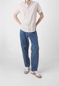 Koszula z krótkim rękawem w beżowe i białe paski, zestawiona z luźnymi niebieskimi jeansami i białymi tenisówkami z czerwonymi akcentami.