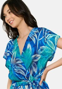 Mėlynas ir žalias tropikų rašto ilgas suknelė su trumpomis rankovėmis ir raišteliu juosmens srityje, puikuojantis dideliais lapų raštais ir v-įpjova.