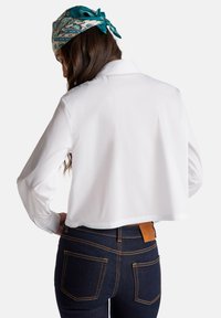 Maglietta bianca a maniche lunghe e taglio corto, vista posteriore. Abbinata a jeans blu scuro con cuciture arancioni e un'etichetta in pelle marrone.