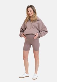 Přetáhnutá béžová mikina na zip s vysokým límcem, v kombinaci s přiléhavými béžovými cyklistickými šortkami. Bílá teniska dokončuje outfit.