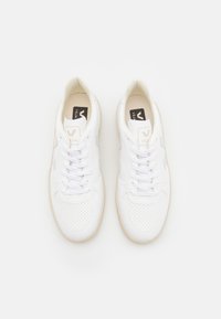 Hvite sneakers med perforert lær, flate lisser og brune gummisåler. Har en logo på tungen og en V-formet aksent på siden.