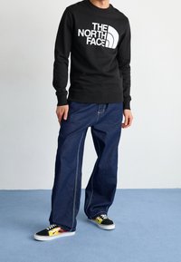 Svart sweatshirt med vitt logo, lösa blå denimbyxor och mångfärgade sneakers med flamdesign på sidan.