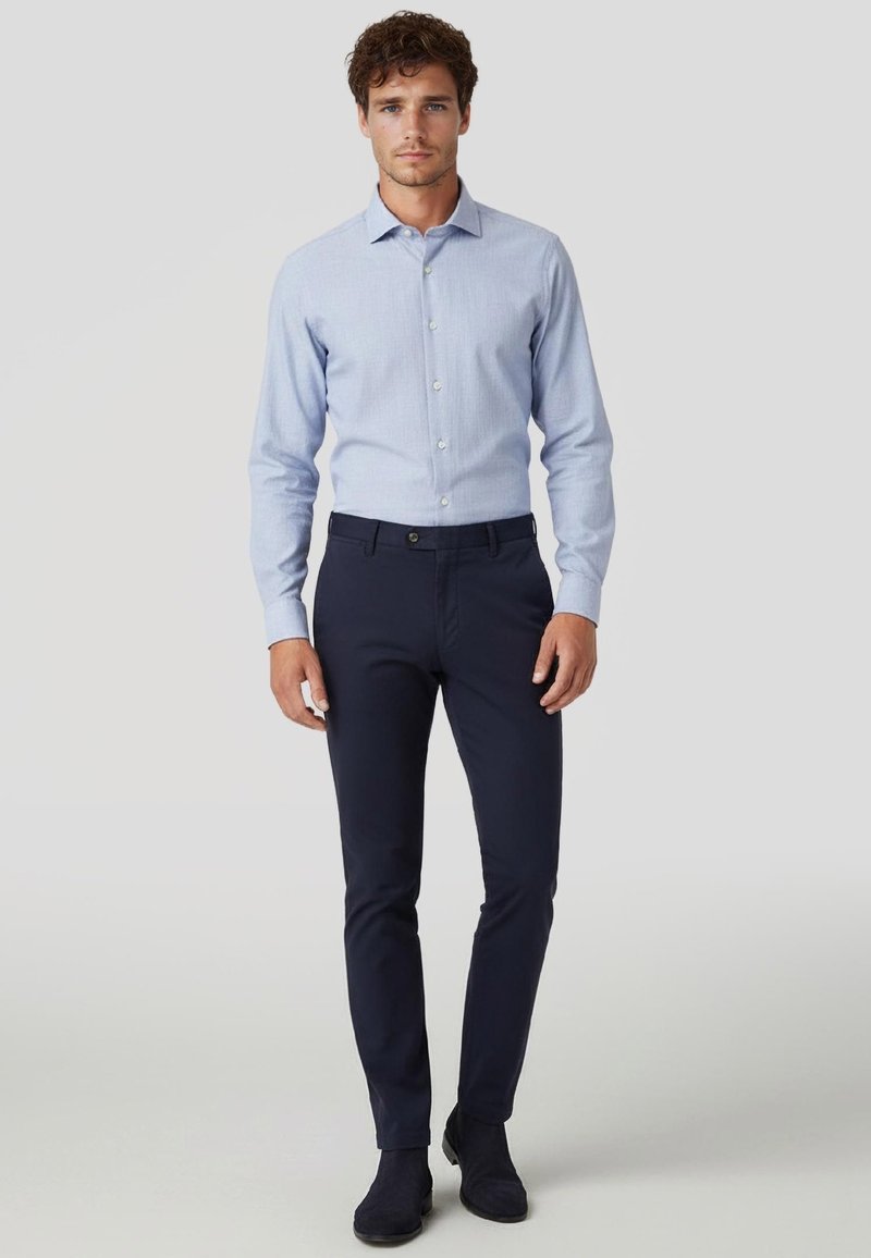 Man staat en draagt een lichtblauw overhemd met lange mouwen, strakzittende donkerblauwe broek en zwarte suède schoenen.