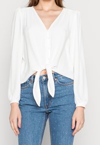 Femme portant un chemisier blanc à manches longues boutonné, noué à la taille, et un jean taille haute bleu à petits pois blancs.
