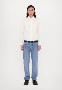 White button-up shirt, blue jeans, black belt, black shoes. Model stands with a neutral pose against a plain white background.