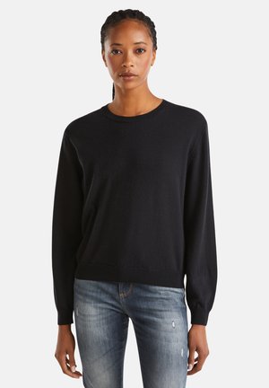 Femme se tenant debout, portant un pull noir à manches longues et un jean déchiré bleu clair, sur un fond uni.