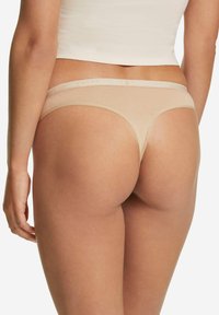 Nude-farbener Tanga mit glatter Textur und elastischem Bund, auf dem der Markenname zu sehen ist. Minimale Rückseitenabdeckung mit schlichtem Design.