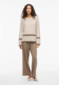 Pull beige à manches longues avec des rayures horizontales marron, accompagné d'un pantalon large taupe et de talons pointus beige clair.