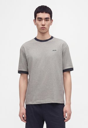 Jeune homme portant un T-shirt gris à manches courtes avec des finitions noires et le logo "BOSS" sur la poitrine, debout devant un fond clair uni.