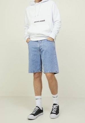Personne portant un sweat à capuche blanc Jack & Jones, un short en denim bleu clair, des chaussettes blanches de marque et des baskets noires sur fond uni.