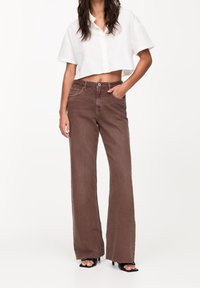 Jeans marron à jambes larges en coton durable avec une taille haute, des poches avant et une texture légèrement délavée. Assortis à une chemise blanche à boutons courte.