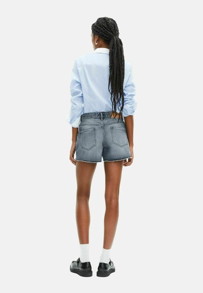 Femme aux longs cheveux tressés portant une chemise bleu clair, un short en denim effiloché, des chaussettes blanches et des chaussures noires, se tenant de dos.