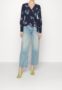 Top floreale blu navy avvolgente con maniche lunghe, abbinato a jeans a gamba larga azzurri e sandali verde con cinturini. Tessuto morbido e dettaglio con fiocco.