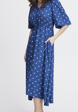 Niebieska sukienka midi w białe kropki, z dekoltem w serek, krótkimi bufkami, paskiem w talii oraz boczną kieszenią, noszona przez stojącą osobę.