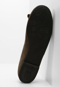 Semelle d'une chaussure plate marron avec motifs en relief de chaussons de ballet et nom de marque sur fond blanc, inclinée contre un mur.
