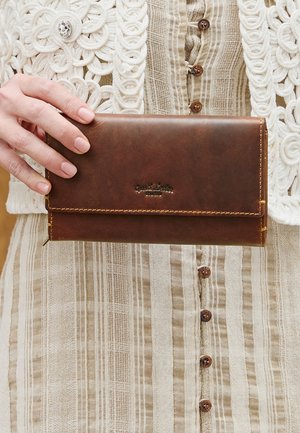 Hand som håller en brun läderplånbok över en beige randig klänning med knappar och en vit virkad cardigan med dekorativ gemknapp.