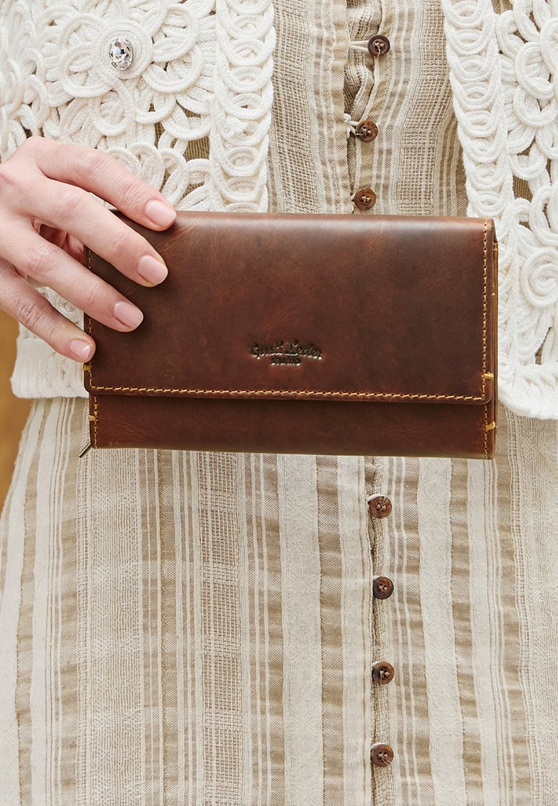 Mano sosteniendo una billetera de cuero marrón sobre un vestido a rayas beige con botones y un chaleco de punto blanco con botón decorativo de joya.