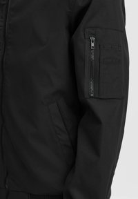 Chaqueta bomber negra con un acabado suave y mate. Cuenta con un bolsillo con cremallera en la manga y bolsillos laterales, puños elásticos y una cintura de canalé.