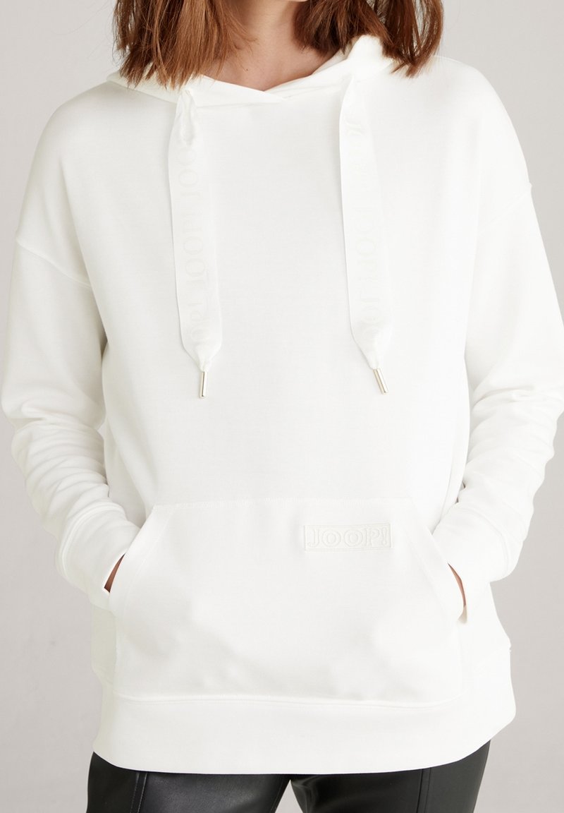 Femme portant un sweat à capuche blanc avec poche avant et cordons, mains dans les poches, associée à un pantalon noir.
