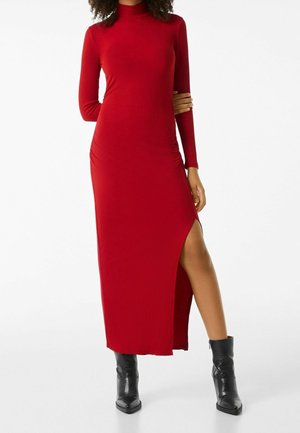 Femme portant une longue robe rouge à col roulé avec une fente latérale haute et des bottines noires, debout avec un bras plié derrière le dos.