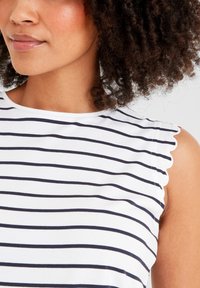Woman with curly hair wearing a white sleeveless shirt with black horizontal stripes and scalloped edges on the sleeves.
