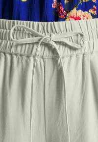 Jasnozielony pasek ściągający z elastycznym wykończeniem, teksturowana tkanina i luźny krój; wzorzysty niebieski element odzieży częściowo widoczny powyżej.