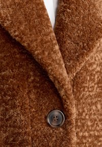 Braun strukturiertes Mantelgewebe mit einer weichen Oberfläche, ausgestattet mit einem großen silbernen Knopf und einem Schalkragen. Nahaufnahme zeigt die Materialdetails.