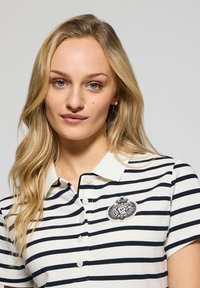 Gestreiftes Poloshirt in Weiß und Marineblau, mit kurzen Ärmeln, einem kragen und einem runden Logo im linken Brustbereich.