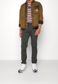 Brun denimjacka med svarta detaljer, randig skjorta, gröna byxor och grå sportskor med neondetaljer. Framsidan, stående pose.