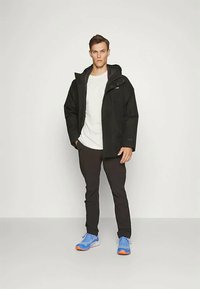 Μαύρο αδιάβροχο μπουφάν με κουκούλα, γκρι επένδυση και ρυθμιζόμενους καρπούς, φορεμένο πάνω από ένα λευκό μπλουζάκι και μαύρο παντελόνι, σε συνδυασμό με μπλε αθλητικά παπούτσια.