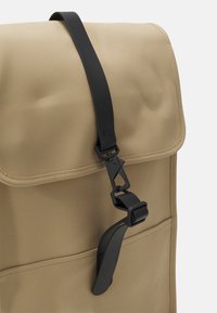 Beige vattentålig ryggsäck med vikbar topp, utrustad med ett svart spännband och en framficka med sömnadsdetaljer.