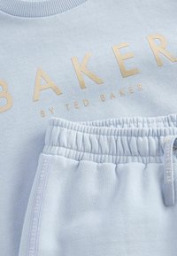 Világoskék pulóver arany betűzéssel, amelyen az "BAKER BY TED BAKER" felirat található, valamint azonos színű melegítőnadrág, elasztikus derékkal és diszkrét márkajelzésekkel.